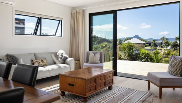 A modern living room with a large sliding glass door leading to a balcony with a scenic view. - Spacious bach close to the beach (Cooks Beach)