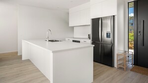 A modern kitchen with white cabinetry, a stainless steel refrigerator, and a built-in island with a sink. - Spacious bach close to the beach (Cooks Beach)