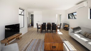A modern living room with a flat-screen TV, wooden coffee table, sofa, and dining area with chairs and a table. - Spacious bach close to the beach (Cooks Beach)