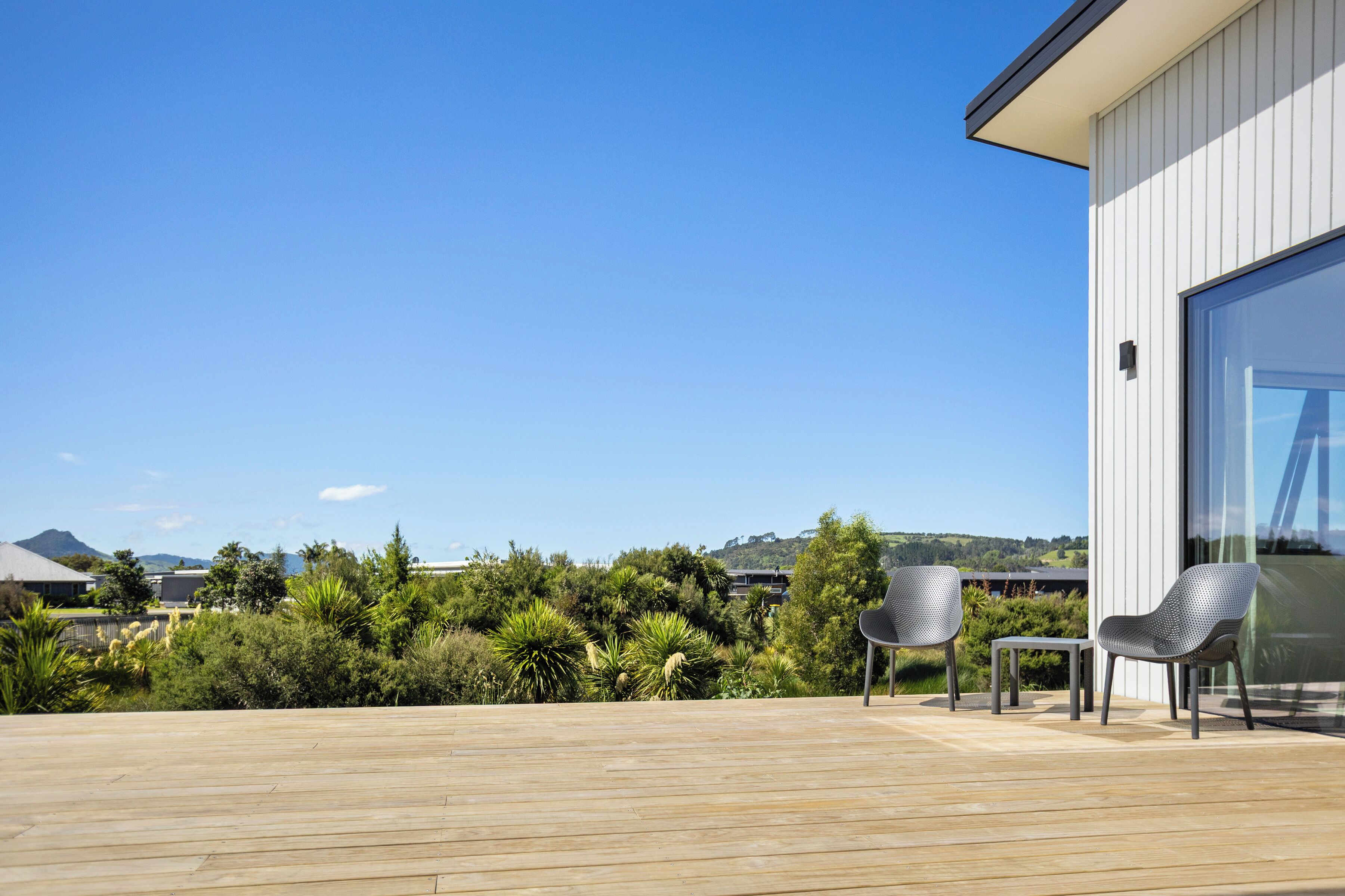 Um deck de madeira com duas cadeiras e uma mesa, com vista para um jardim exuberante e um edifício moderno com grandes janelas.