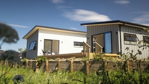 A modern two-story house with a flat roof, large windows, and a wooden deck. - Spacious bach close to the beach (Cooks Beach)