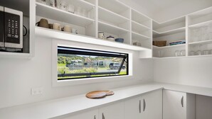 A modern kitchen with white cabinetry, a built-in microwave, and a window with a view of a residential area. - Spacious bach close to the beach (Cooks Beach)