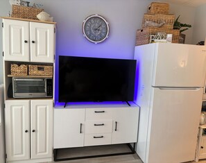 Una cocina con armarios blancos, un televisor de pantalla plana sobre un soporte, un microondas, un refrigerador y un reloj de pared.