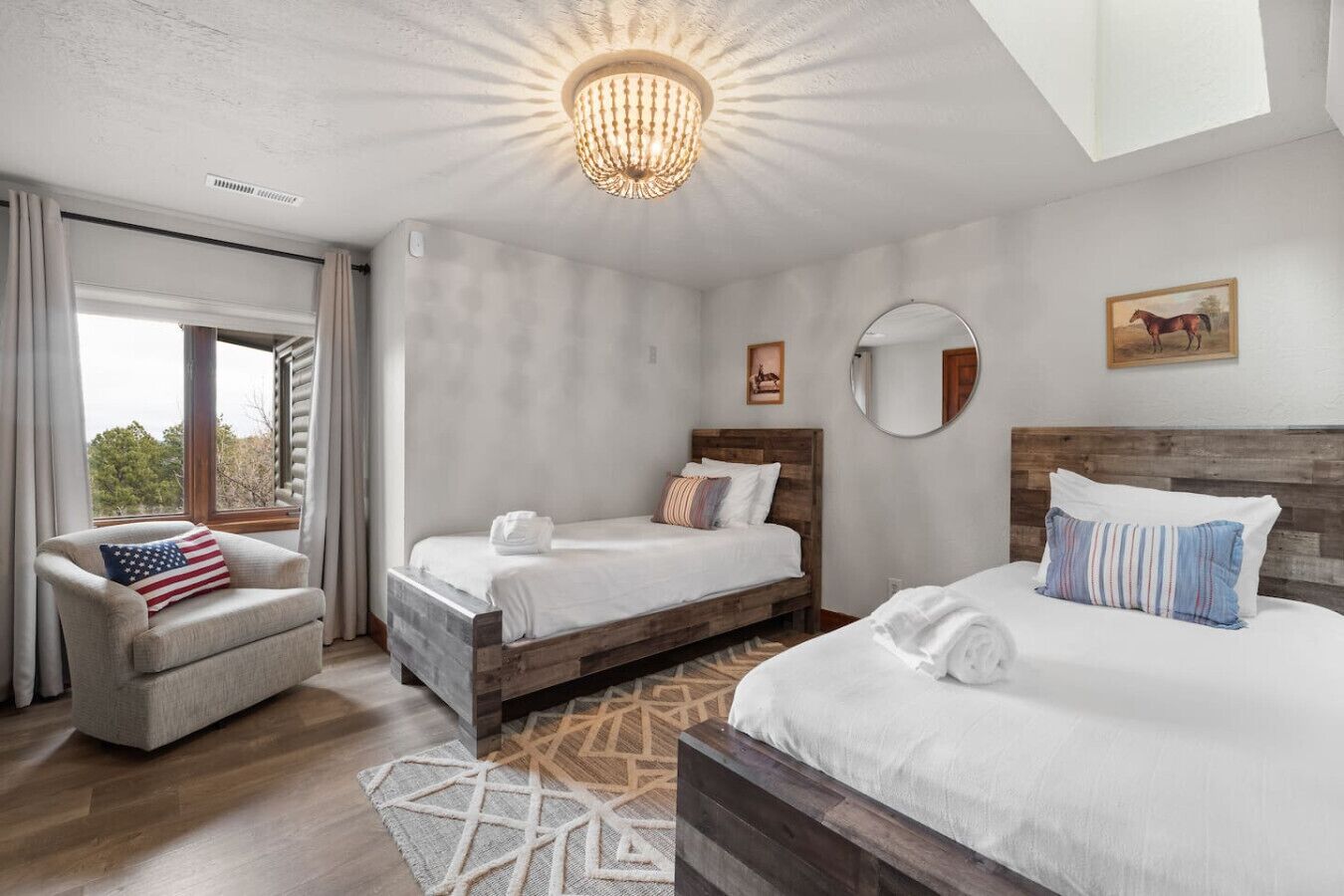 A bedroom with two beds, a chair, a chandelier, and a large window.