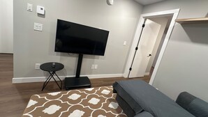 Un salon avec un téléviseur à écran plat posé sur un meuble, une petite table ronde, un tapis à motifs et un canapé gris.