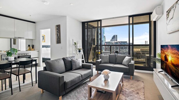 Un appartement moderne comprenant une cuisine, deux canapés gris, une table basse et une grande fenêtre donnant sur la ville.