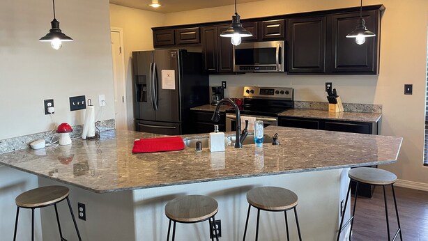 A modern kitchen with dark cabinetry, a granite countertop, a stainless steel refrigerator, a microwave, and a gas stove. There are three bar stools and pendant lights above the island.