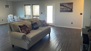 A spacious living room with a beige sofa, a dining table with chairs, a ceiling fan, and a cityscape painting on the wall.