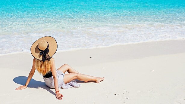 Eine Frau sitzt an einem Strand, trägt einen Hut und blickt auf ein ruhiges Meer und eine kleine Insel in der Ferne.