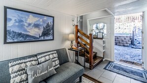 A cozy living room with a gray sofa, a framed landscape painting, and a wooden staircase leading to a door. - Mountain Top House with a View - Elk House (Ruidoso)