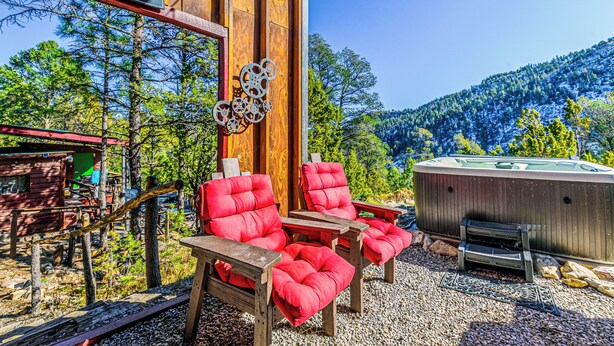 A rustic outdoor seating area with red cushioned chairs and a hot tub, surrounded by wooden structures and a mountainous backdrop. - Mountain Top House with a View - Elk House (Ruidoso)