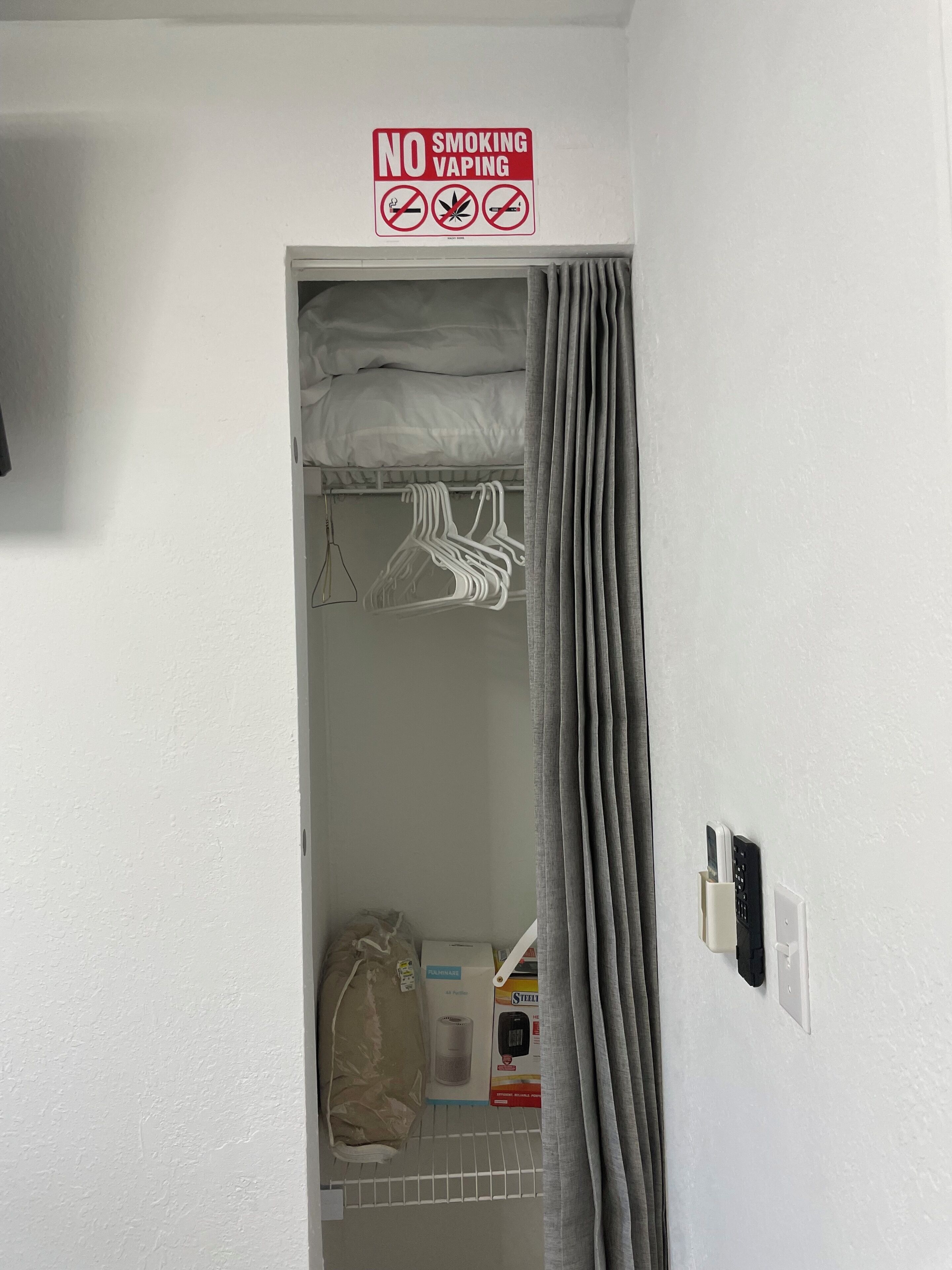 Een hotelkamer met een kast met hangende kleding en een plank met toiletartikelen. Een 'Niet roken' bordje is boven de kast te zien.