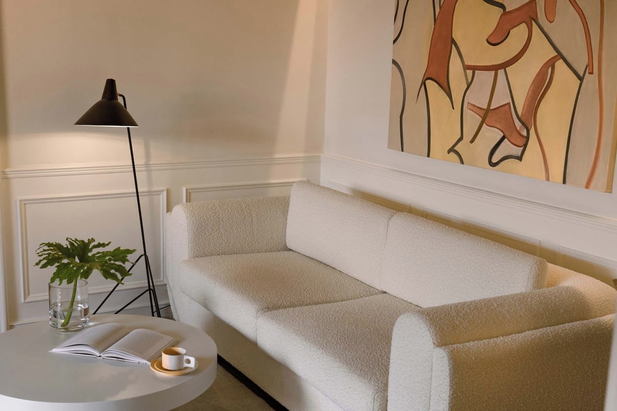 A living room with a white sofa, a round coffee table with a book and a cup, a floor lamp, and a wall with abstract art.