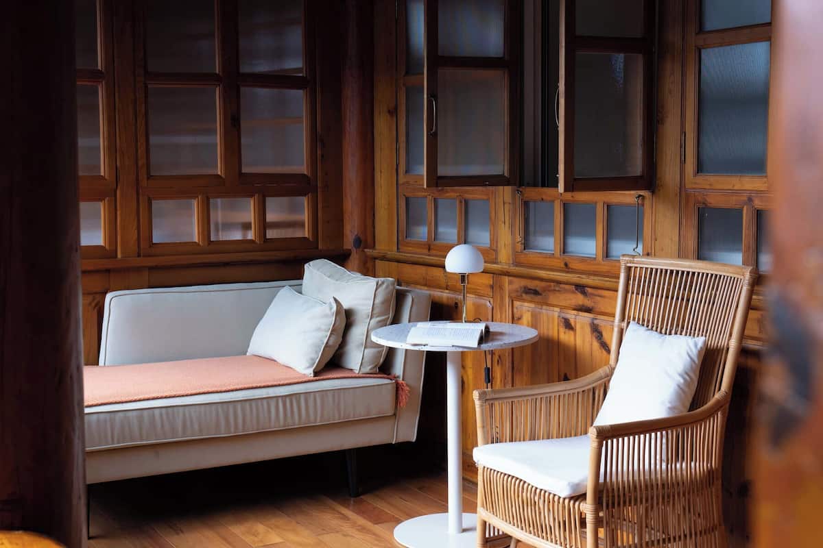 A cozy room with a wooden paneled wall, a white sofa with cushions, a wicker chair, and a small round table with a lamp.