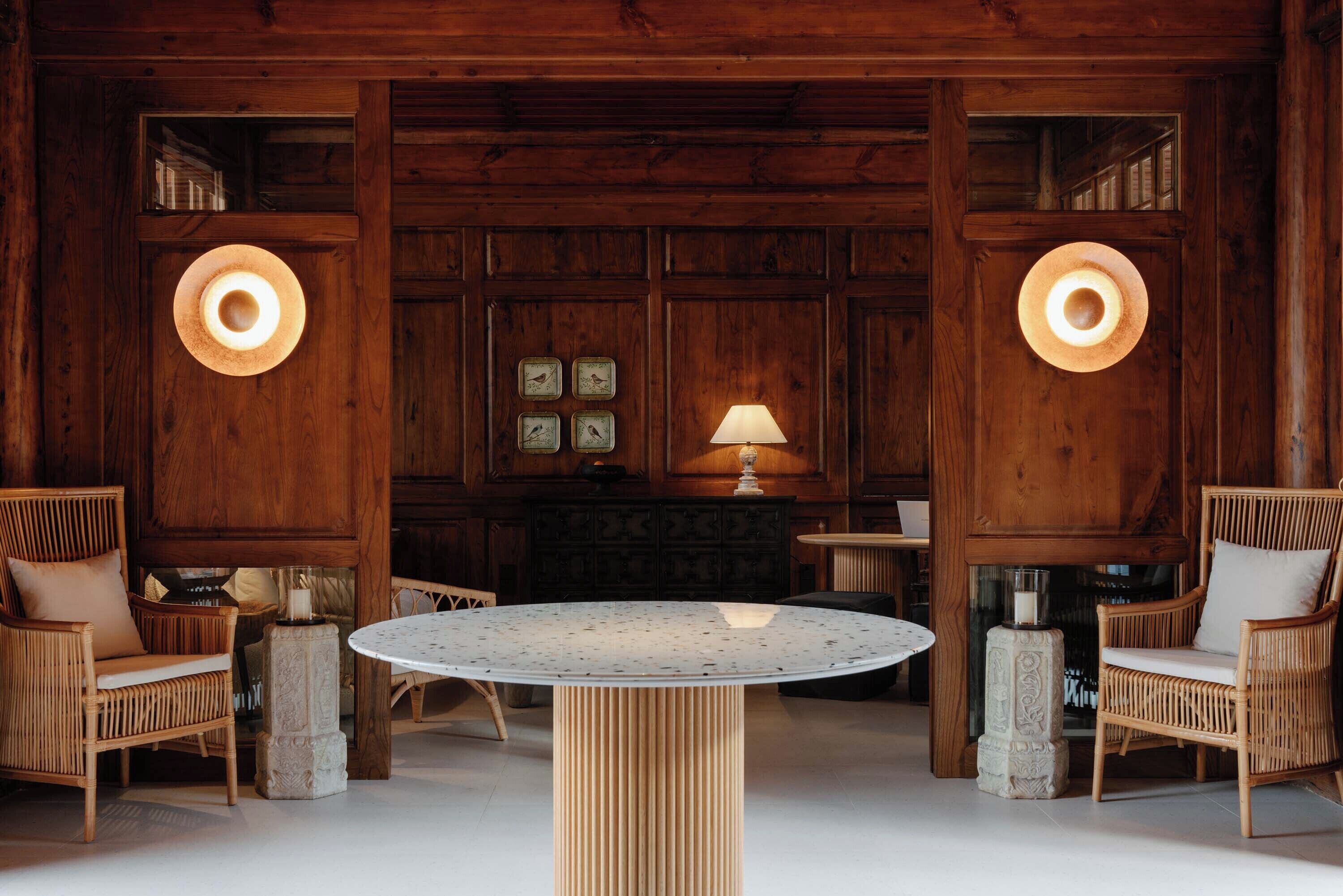 Un intérieur en bois avec une table ronde, des chaises en osier et des lampes fixées au mur.