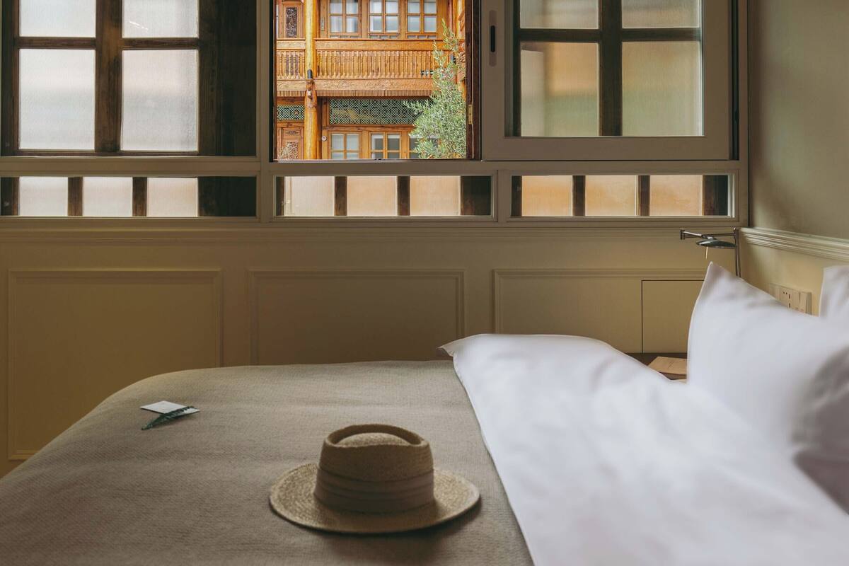 A hotel room with a bed, a hat, and a window with a view of a building.