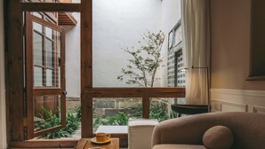 Ein gemütliches Zimmer mit Fensterrahmen aus Holz, einer Couch, einem Tisch mit Buch und Tasse sowie Blick ins Grüne.