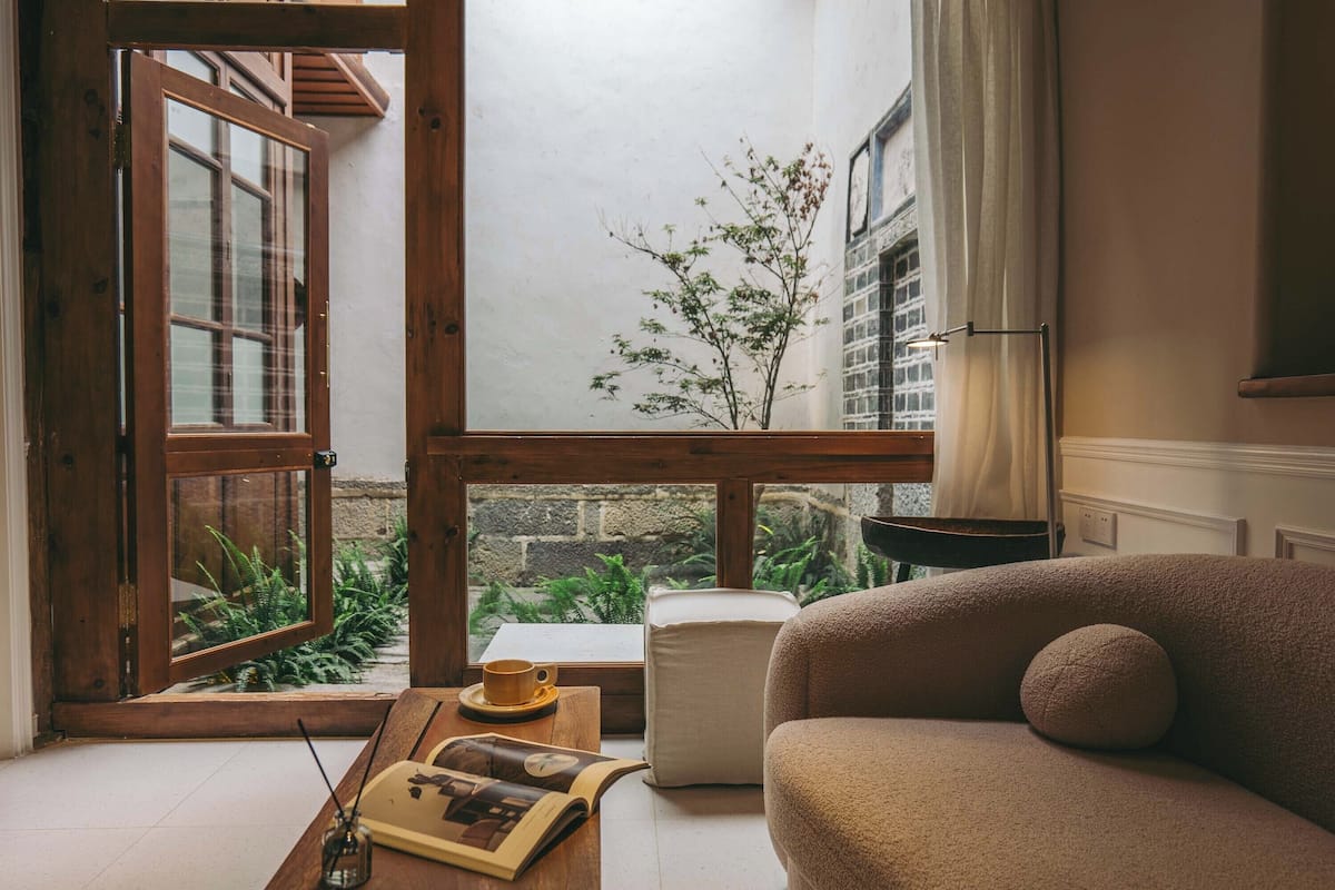 A cozy room with a wooden-framed window, a sofa, a table with a book and a cup, and a view of greenery outside.