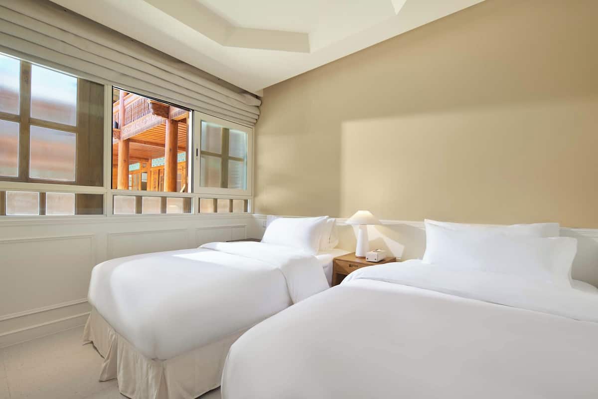 A hotel room with two beds, a window with blinds, a nightstand with a lamp, and a view of a wooden structure outside.