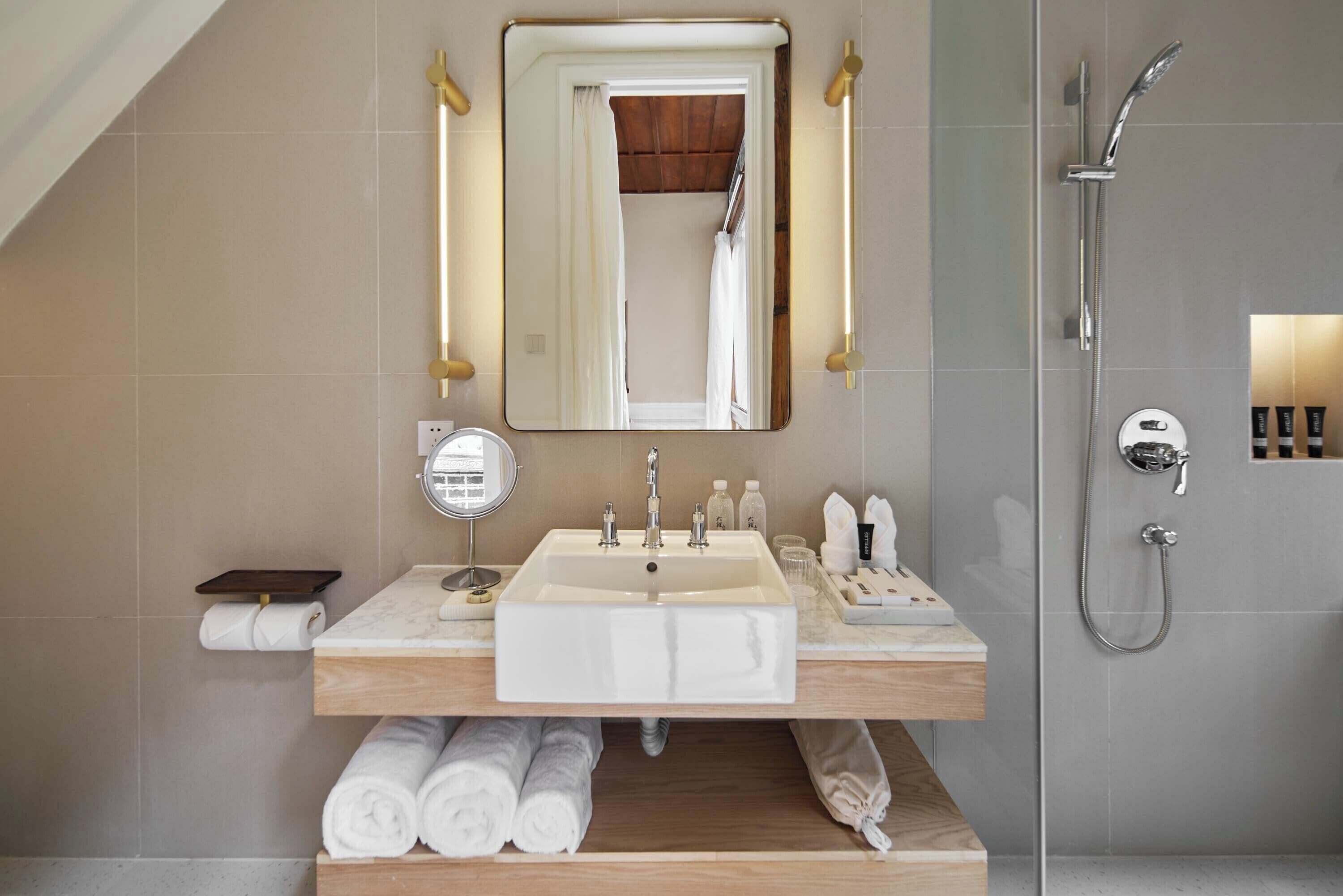 A modern bathroom with a large mirror, two sinks, a shower, and neatly rolled towels.
