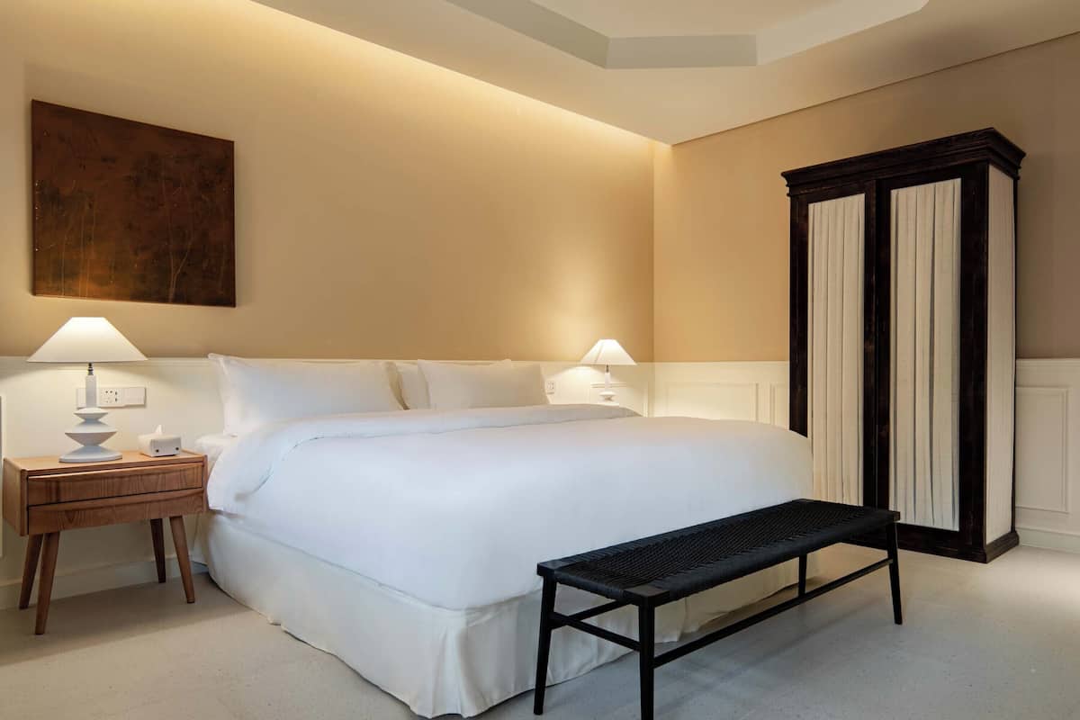 A neatly arranged bedroom with a large bed, bedside lamps, a wooden nightstand, a bench, and a dark wooden wardrobe.
