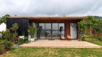 A modern house with a thatched roof, large glass windows, and a patio with two chairs.