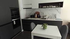 Una cocina moderna con armarios blancos, salpicadero de mármol y horno empotrado. Hay una mesa de comedor con dos sillas y una planta en maceta.