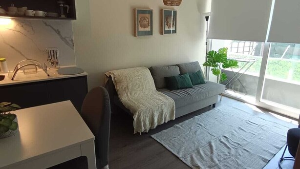 Un moderno salón con un sofá gris, una mesa de centro blanca y un comedor con una mesa y sillas blancas. Una ventana grande ofrece vistas a zonas verdes.