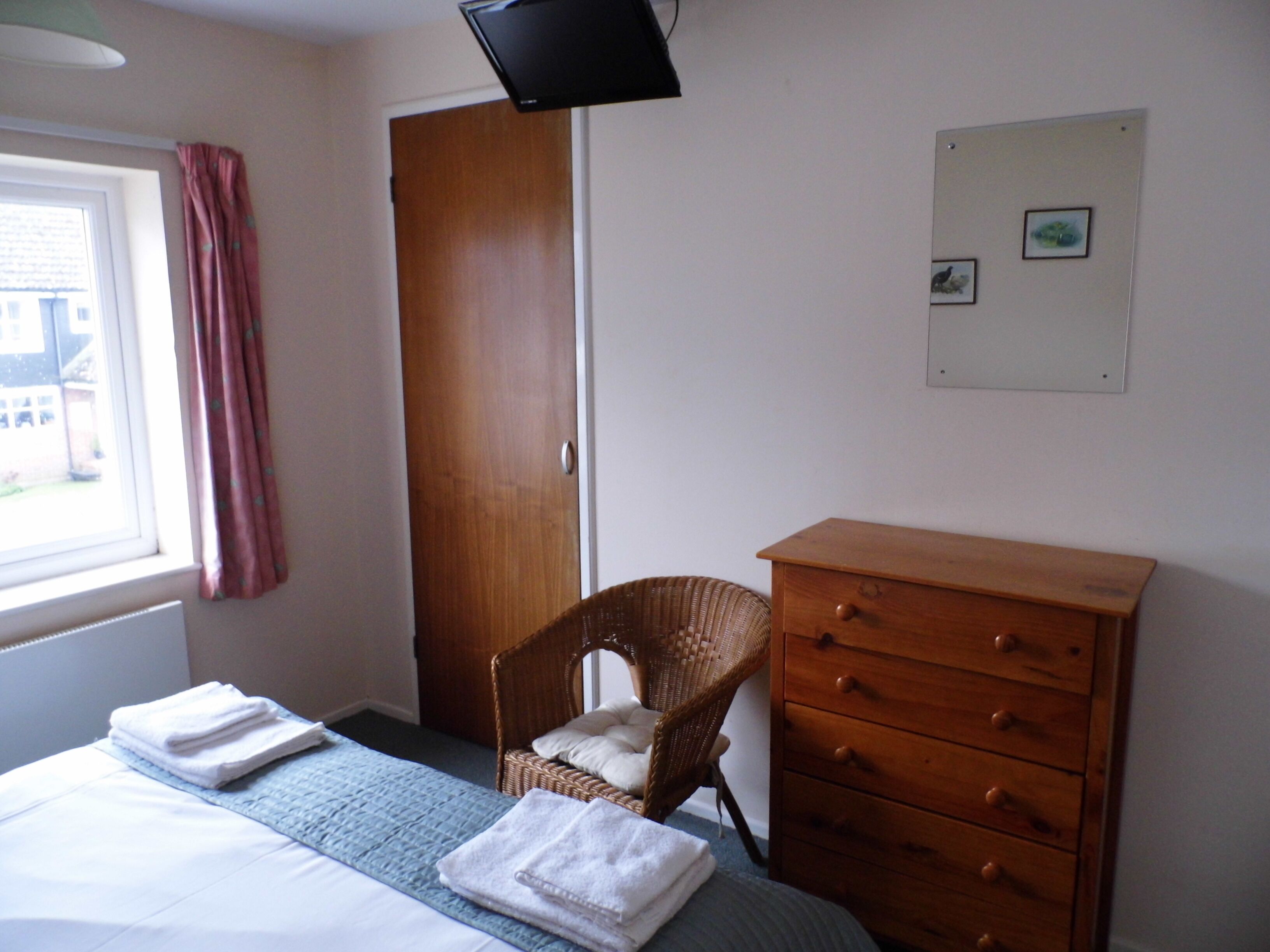 Una camera da letto con un letto, una cassettiera in legno, una sedia in vimini, una finestra con tende e un quadro appeso al muro.