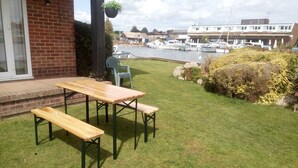 A patio with a wooden table and benches, a brick building, and a marina in the background. - Wherry, pet friendly cottage. on the riverside with free fishing and mooring. (Wroxham)