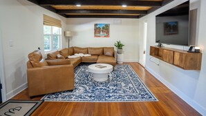 A living room with a sectional sofa, a round coffee table, a patterned rug, a flat-screen TV, and wooden beams on the ceiling. - Minnesota Manor Home & Guest House-Your DeLand Getaway (DeLand)