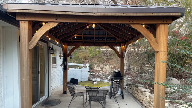 A wooden pergola with a table and chairs, a grill, and string lights.
