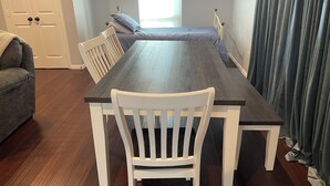 A dining area with a dark wooden table, white chairs, a grey sofa, and a bed with a blue mattress.