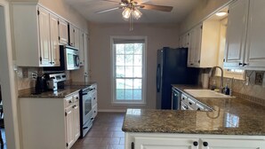 A kitchen with white cabinets, a granite countertop, a built-in oven, a refrigerator, and a window with a view of trees.