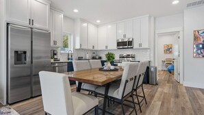 Une cuisine moderne avec des meubles blancs, des appareils en acier inoxydable et une table à manger en bois, accompagnée de chaises.