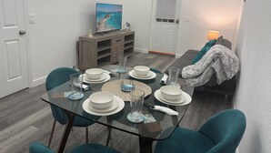A dining area with a glass table set for four, surrounded by teal chairs. A wooden cabinet with a television and decorative items is against the wall.