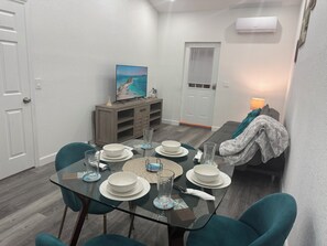 A dining area with a glass table set for four, surrounded by teal chairs. A wooden cabinet with a television and decorative items is against the wall.