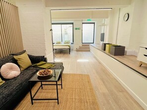 Quarto de hotel moderno com sofá cinza escuro, uma pequena mesa com uma tigela de petiscos, um tapete tecido, uma parede branca e um design de ripas de madeira à esquerda.