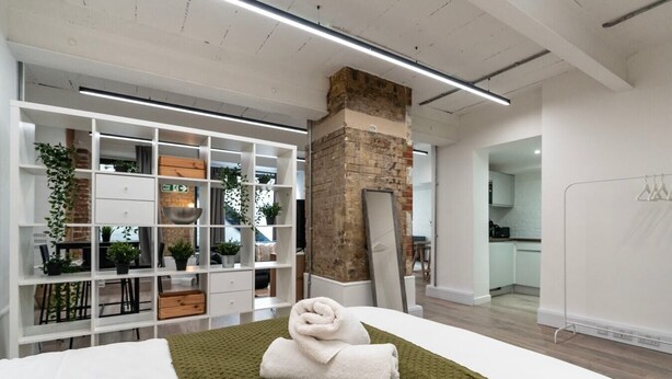 Habitació d'hotel moderna amb un llit gran, un rug texturat verd, parets blanques, una paret decorativa de maó i una prestatgeria blanca amb plantes i objectes decoratius.