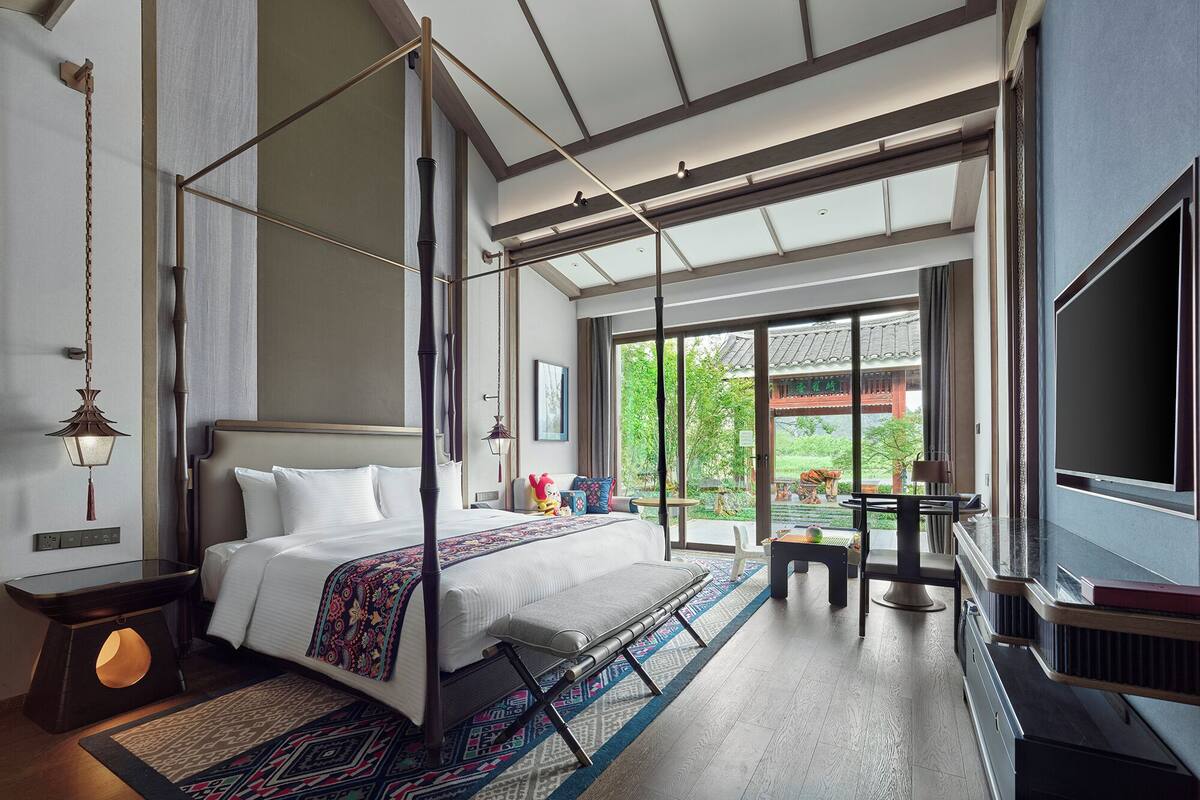 A modern bedroom with a large bed, a TV, and a view of greenery outside.