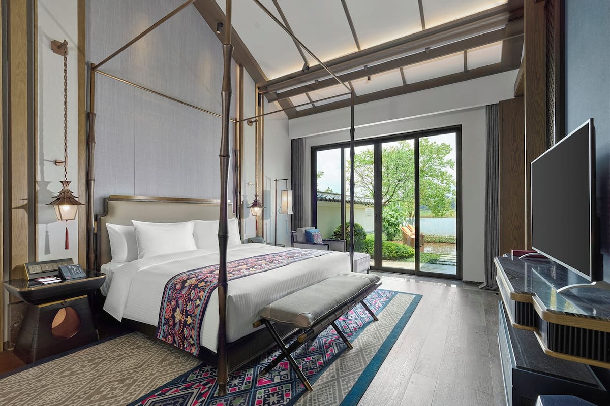A modern hotel room with a four-poster bed, a flat-screen TV, and a large sliding glass door to the outdoors.