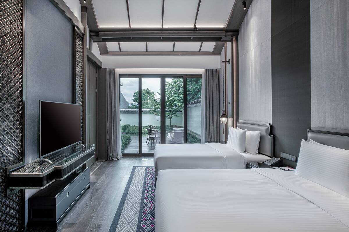 A modern hotel room with a large bed, a flat-screen TV, and a balcony with a view of greenery.