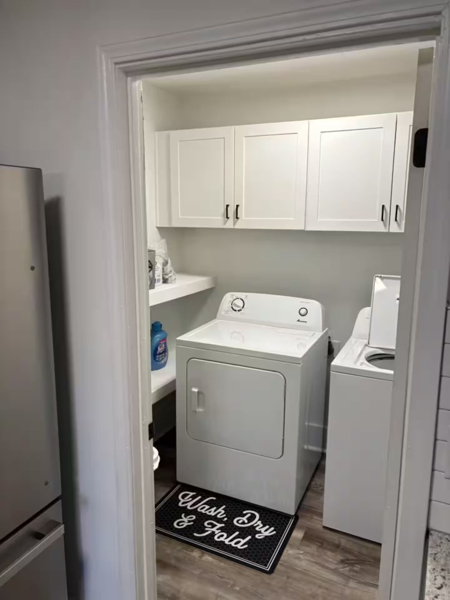 Una sala de lavandería con lavadora y secadora de carga frontal, armarios blancos y un felpudo que dice “Wash, Dry & Fold”.