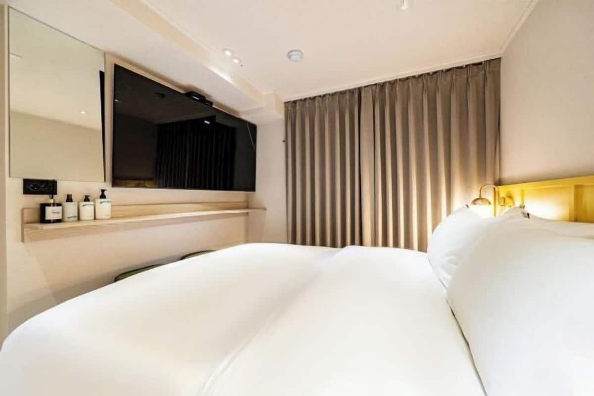 A modern hotel room with a large bed, a bedside table with toiletries, a mirror, and a window with curtains.