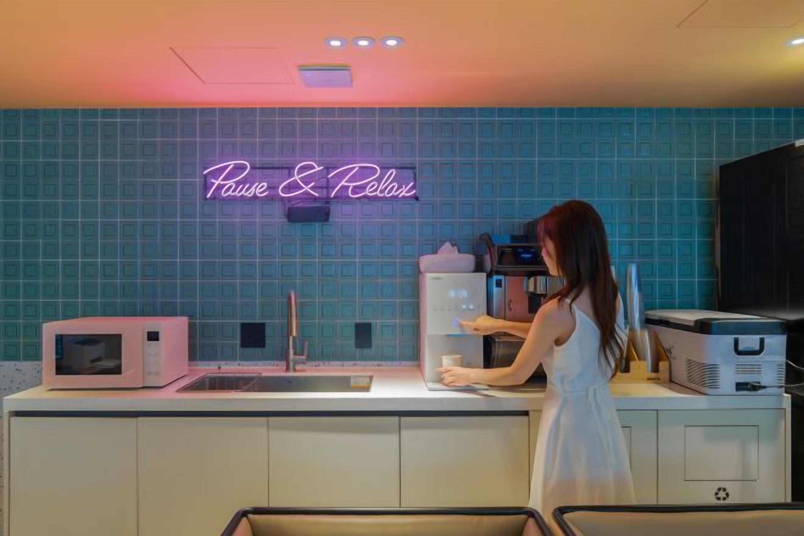 A kitchen with a neon sign reading 'Pause & Relax,' a microwave, a sink, and a person preparing food.