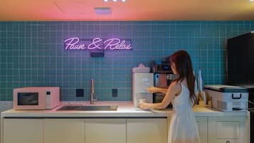 A kitchen with a neon sign reading 'Pause & Relax,' a microwave, a sink, and a person preparing food.