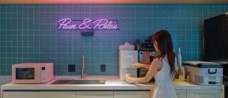 A kitchen with a neon sign reading 'Pause & Relax,' a microwave, a sink, and a person preparing food.