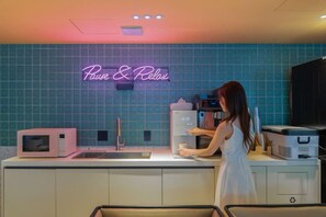A kitchen with a neon sign reading 'Pause & Relax,' a microwave, a sink, and a person preparing food. - Sinchon APL Hotel (Seoul)