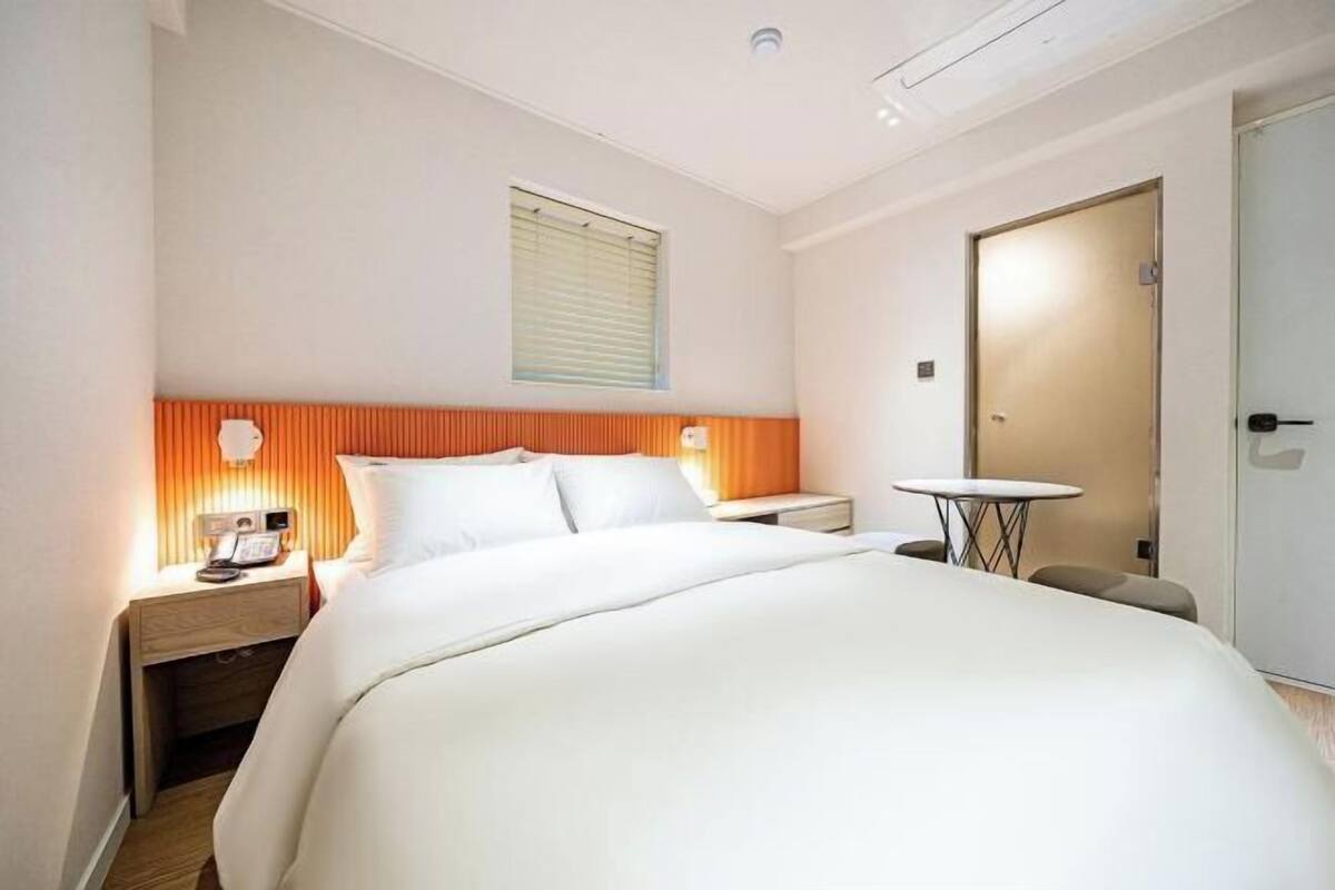 A hotel room with a bed, bedside tables, a small round table, a door, and a window with blinds.