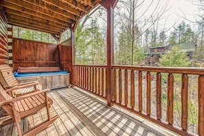 Eine Holzterrasse mit Whirlpool und Sitzgelegenheit, umgeben von Holzgeländer und Bäumen.